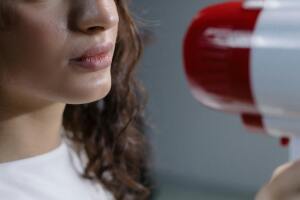 Close-up of a woman speaking into a red and white megaphone against a gray background.
