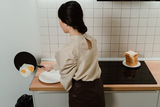 A woman frying eggs in a modern kitchen, captured from behind showing a stack of bread nearby.