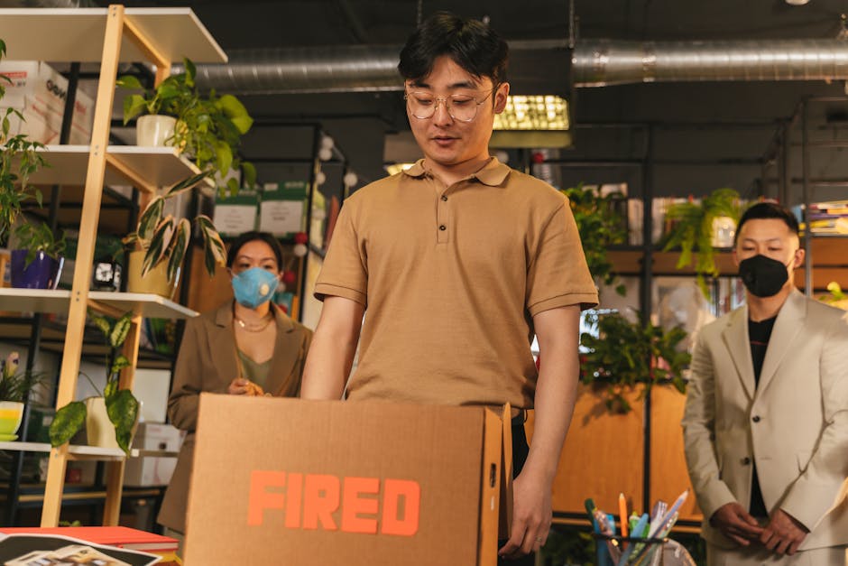 Man standing by cardboard box labeled 'fired', in an office surrounded by colleagues.