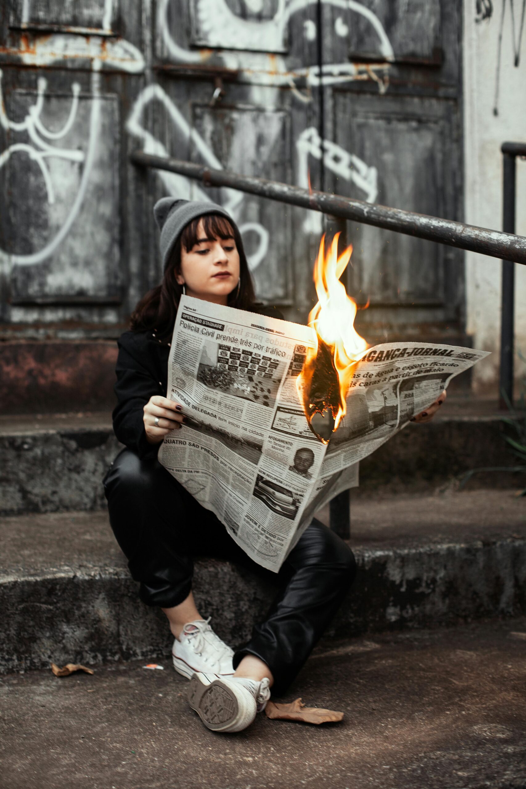 Woman sits outdoors reading a newspaper on fire, creating a dramatic urban scene.